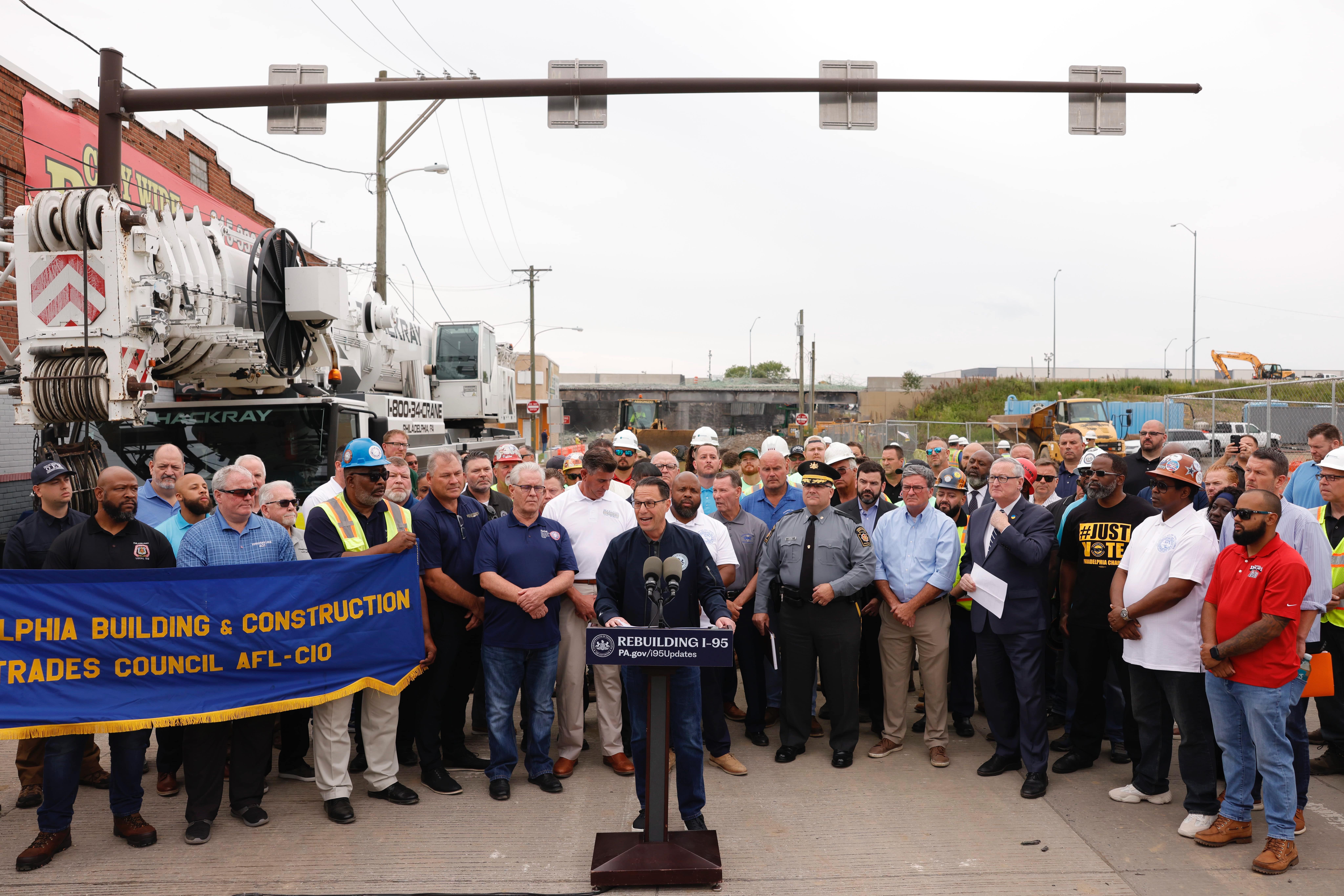 Governor Shapiro stands behind a podium that has a Rebuilding I-95 sign on it, as he talks to reporters during a press conference at the bridge collapse site, while various transportation officials, law enforcement and construction workers stand in the background